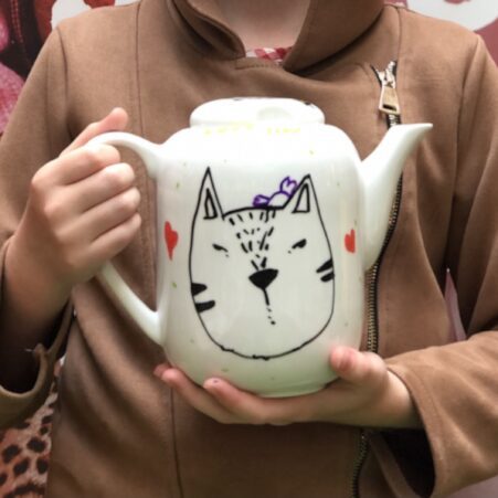 een frontfoto van een kind die een theepot inkleurde met een poezenhoofd met porseleinstiften handgemaakt in het atelier van Hello Mojo in Heule. 18. Milieubewuste activiteiten voor groepen op milieu-evenementen met Hello Mojo